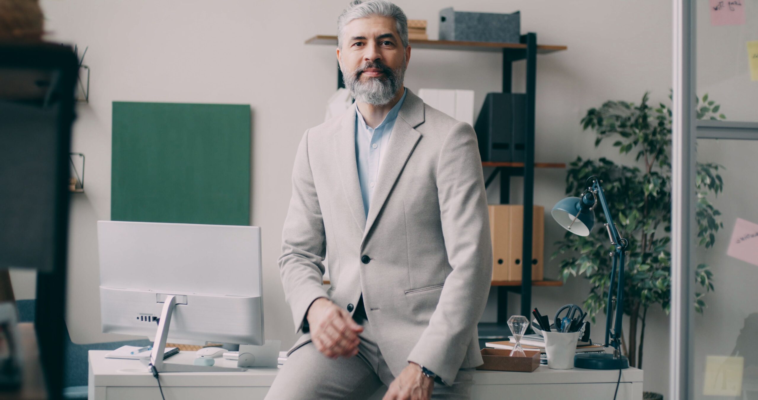 A mature businessman in a beige suit sits confidently on a desk in a modern office setting.