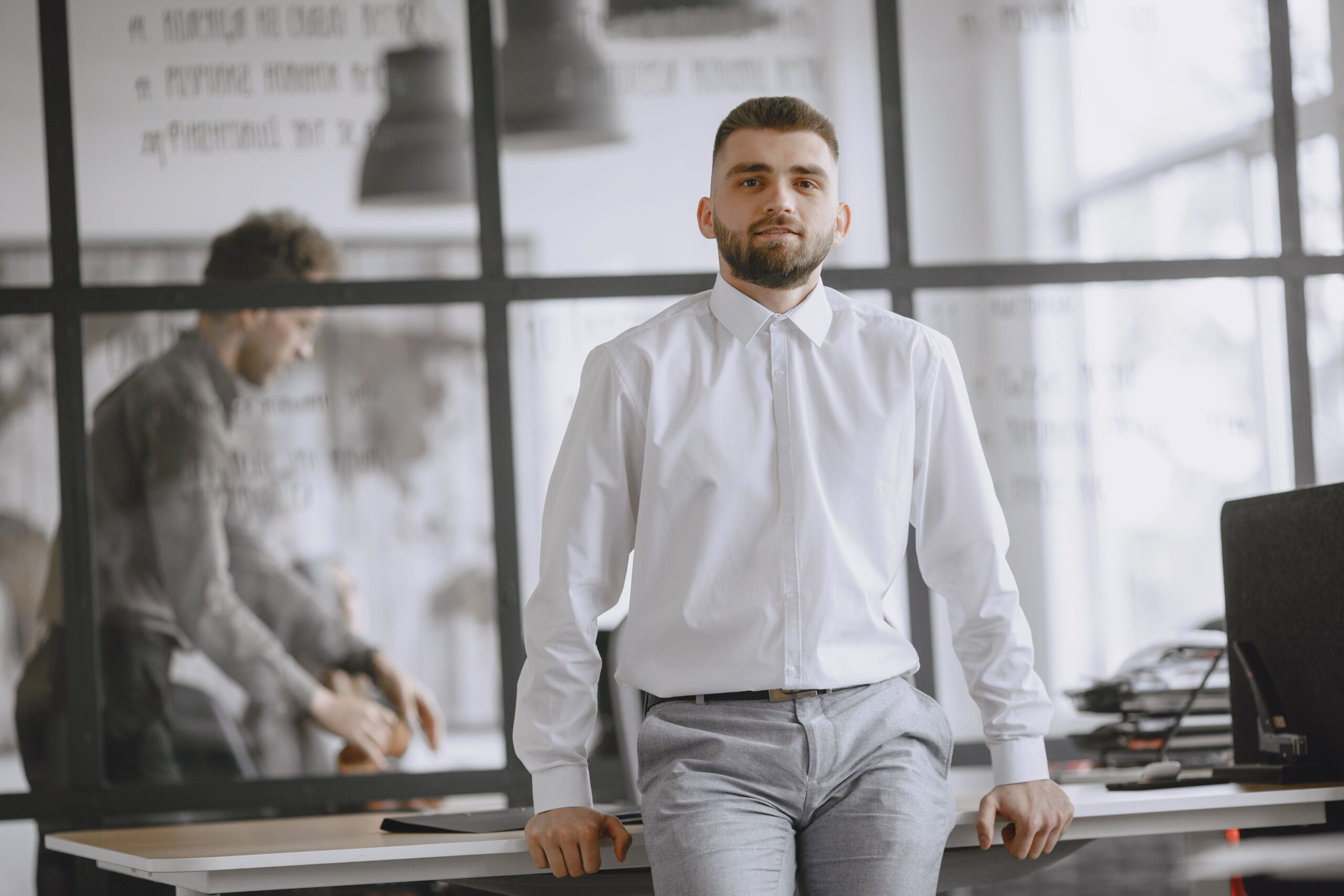 Confident man in white shirt leaning on desk in modern office with glass partitions.